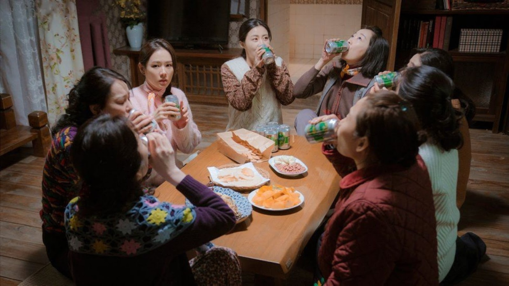 Seri junto al grupo de mujeres del pueblo norcoreano celebrando con cerveza y comida, sentadas en una mesa. Imagen promocional de Crash Landing On You de parte de tvN.