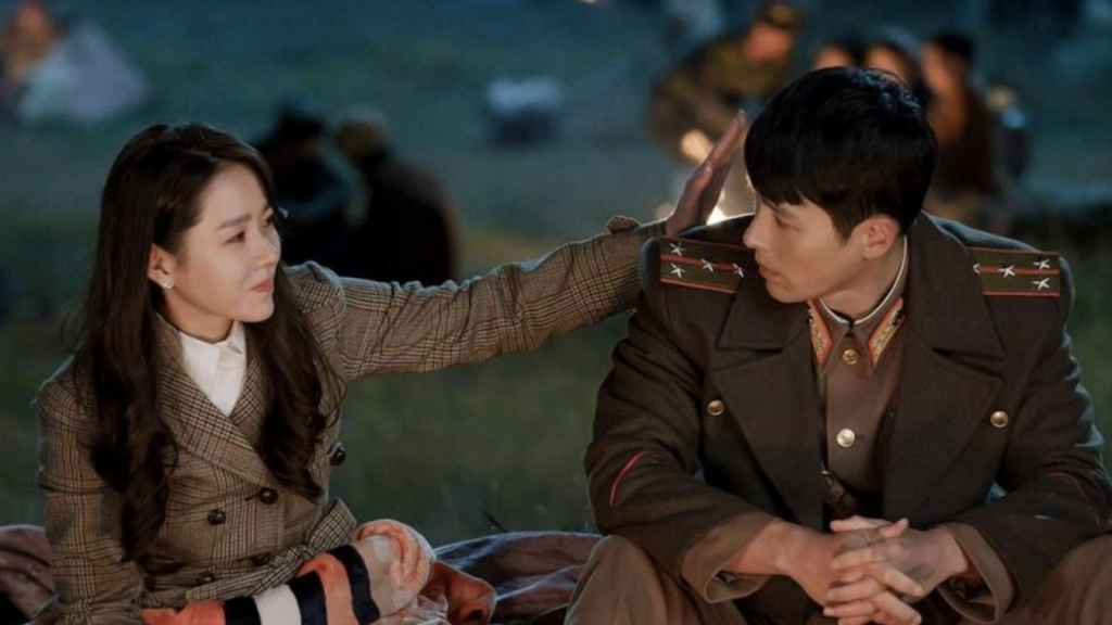 Imagen promocional de Crash Landing on You, donde Seri le toca la cabeza a el capitán Ri sentados en el suelo, de noche, mientras se miran a los ojos. Imagen de parte de tvN.